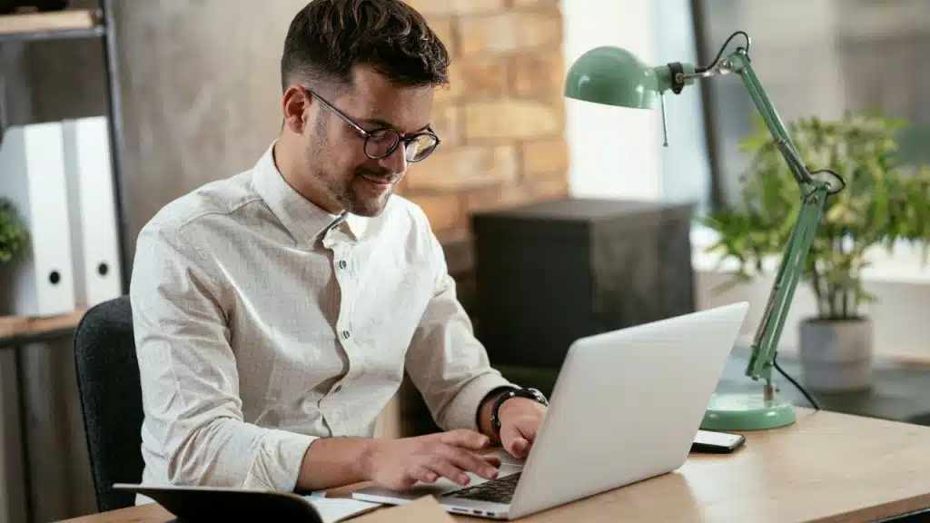 A smiling, bespectacled man in a white collared shirt types on a silver laptop at a desk.