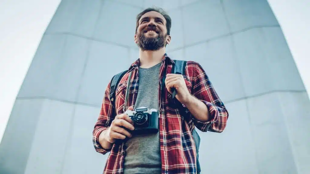 A smiling bearded man in a plaid shirt and backpack holds a vintage camera outdoors.