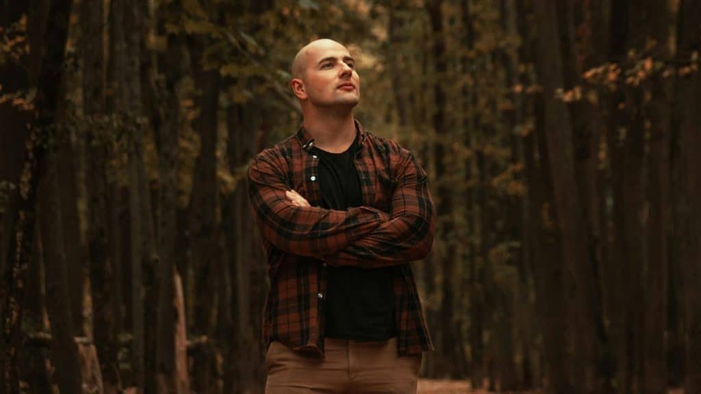 A bald man in a red and black plaid shirt stands in a dark, tree-lined path, arms crossed.