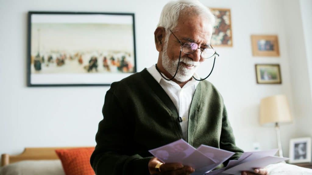 An elderly man with a white beard and glasses smiles while looking down at a stack of photos.