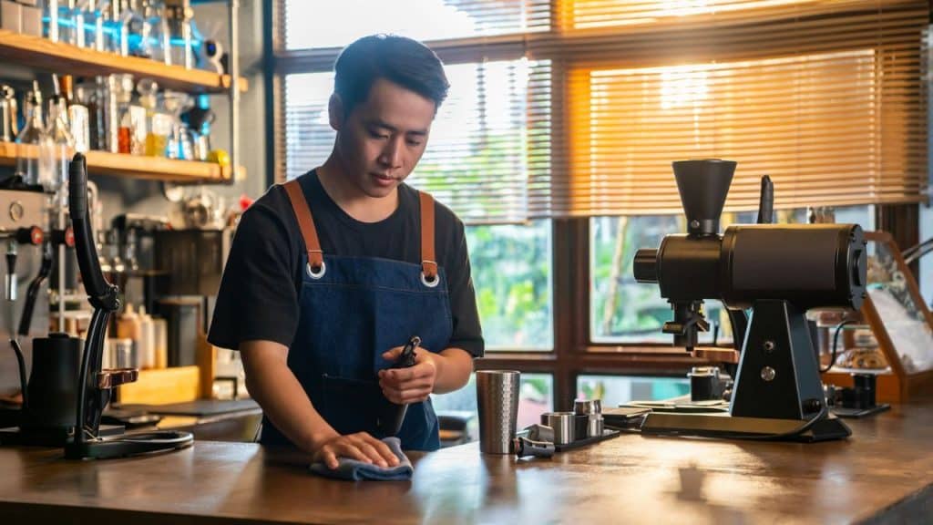 A young barista in a denim apron wipes down a wooden counter next to a coffee grinder.