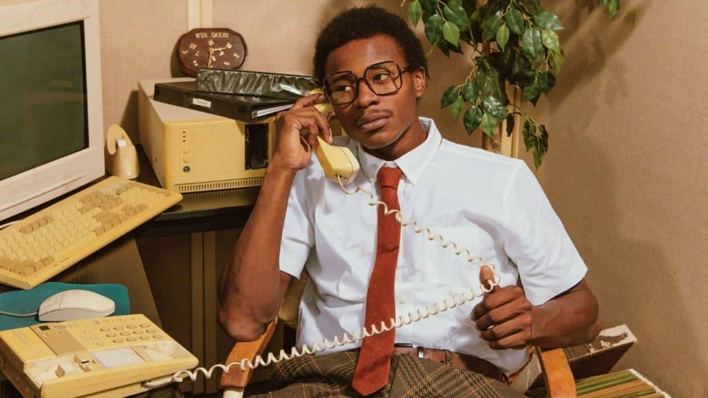 A young man with glasses and a tie talks on a yellow rotary phone in a retro office setting.