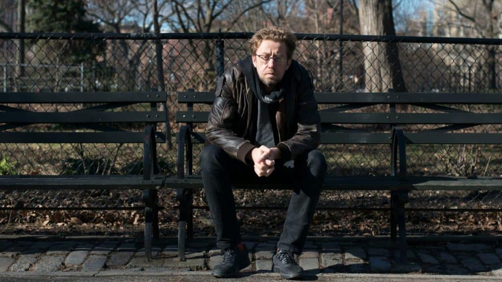 A man with glasses, wearing a leather jacket, sits alone on a dark park bench.