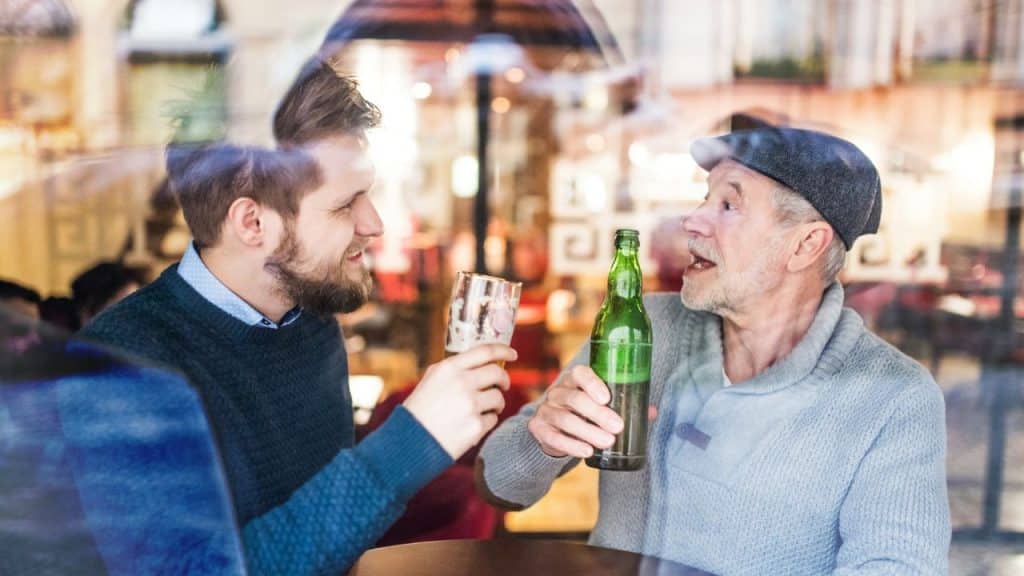 Two men, one young and one older, cheer with beer through a reflection-filled window.