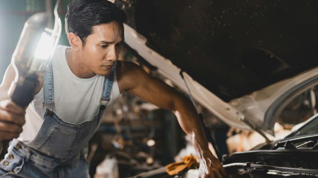 A mechanic in denim overalls holds a flashlight, looking under the hood of a car.