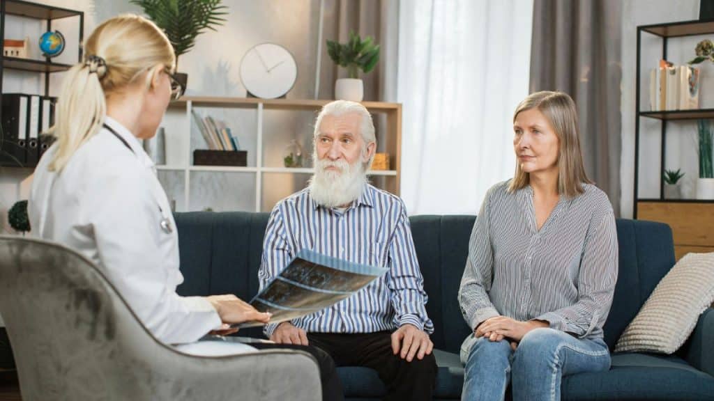 A doctor shows an X-ray to an older bearded man and a woman sitting on a couch.
