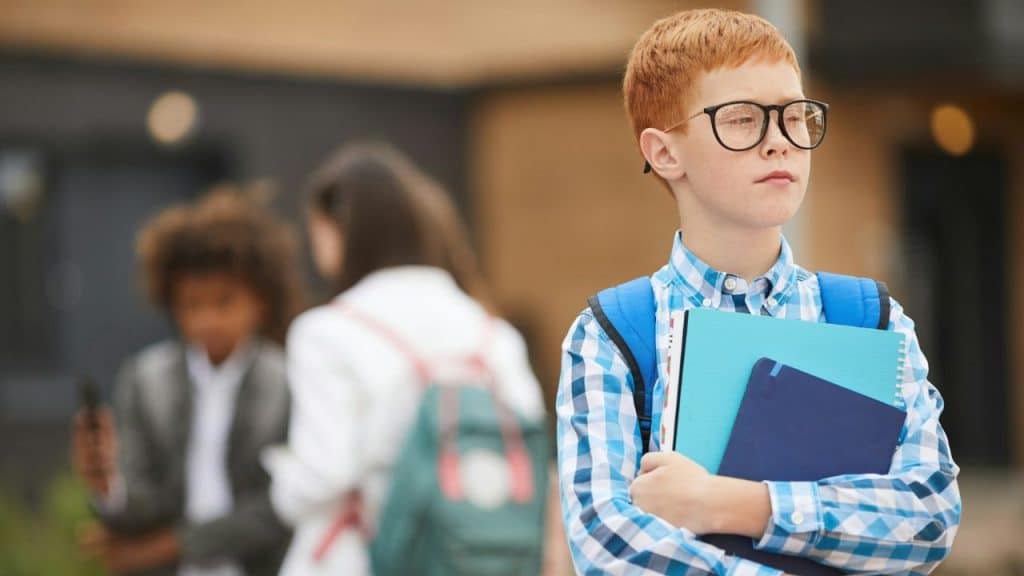 A boy with red hair, glasses, and a backpack holds books outside a school.