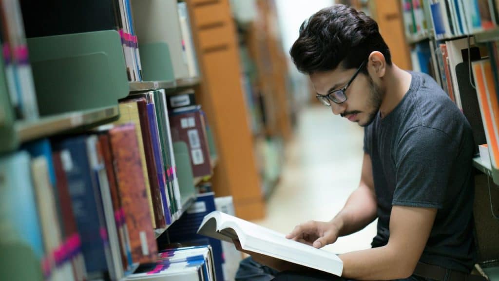 A young man in glasses sits in a library aisle reading a large, open book.