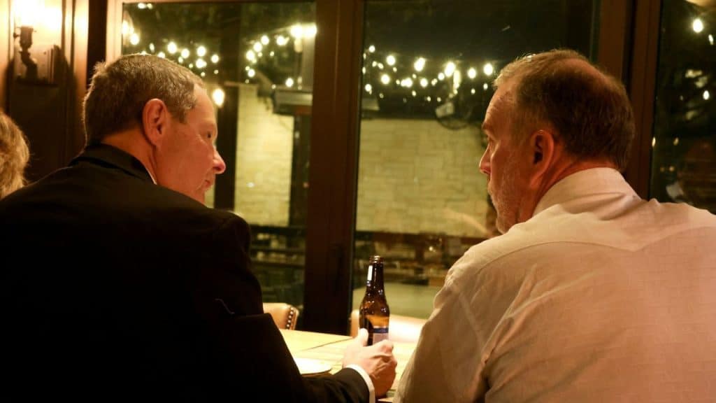 Two men sit at a table facing each other, with a window and lights behind them.
