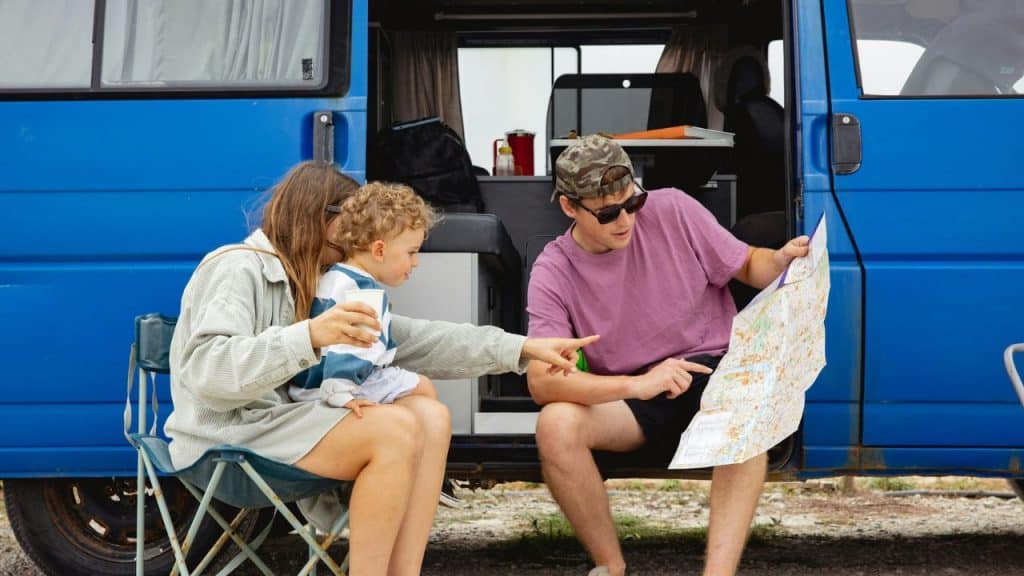 A family sits by their open blue van, looking at a map while outdoors.