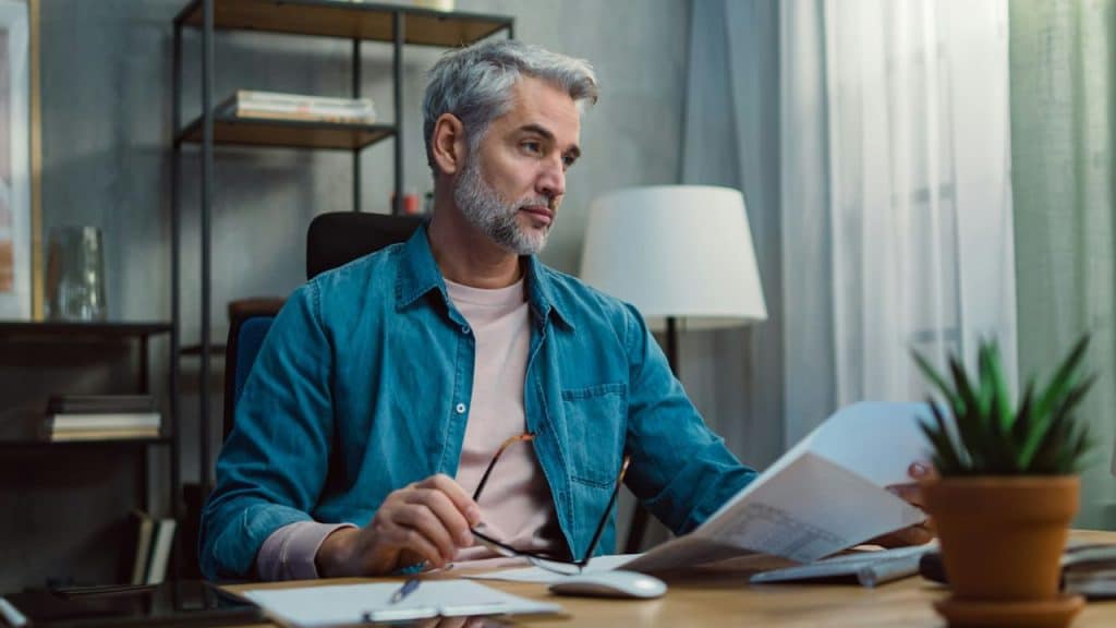 A silver-haired man at a desk is reviewing papers while holding a pair of glasses.