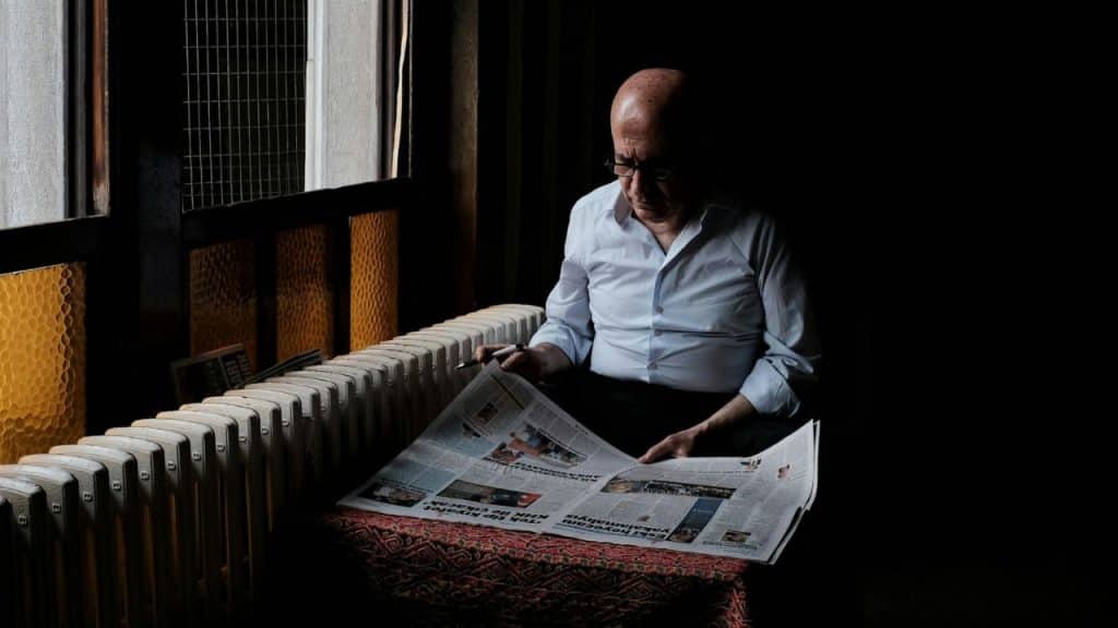 An older man sits in a dark room by a radiator, reading a folded newspaper.