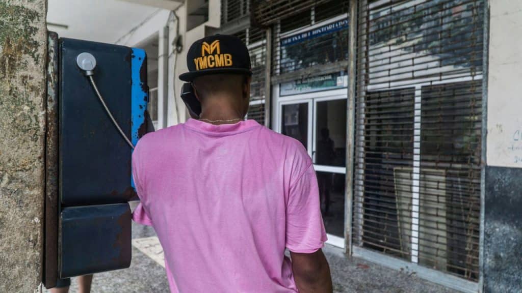 A person in a pink shirt and black hat uses an old payphone outdoors.
