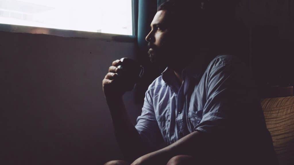 A man sits in a dark room by a window, holding a mug to his mouth.