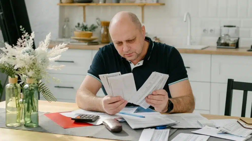 A bald man sits at a kitchen table, intently sorting through a stack of papers.
