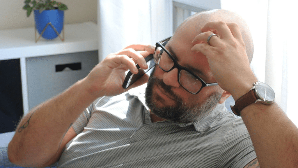 A bearded, bald man with glasses is on a phone call, looking stressed and rubbing his head.
