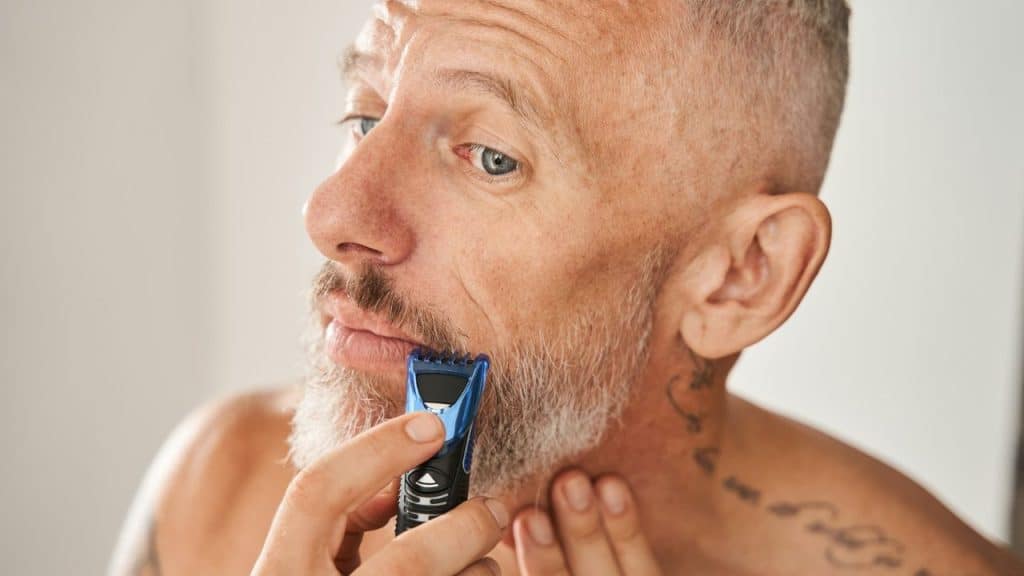 An older man with a faded crew cut trims his gray beard using a handheld electric trimmer.