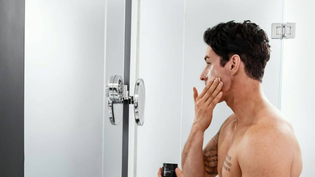 A bare-chested, tattooed man applies a white cream to his cheek in a modern bathroom.