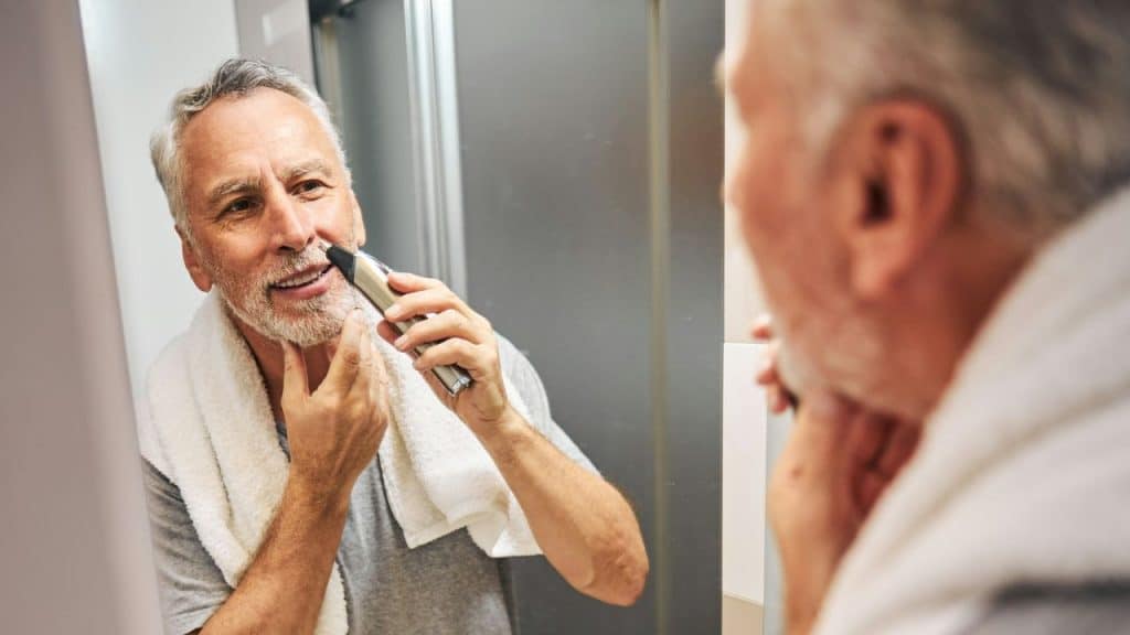 A smiling, gray-haired man in a towel uses a handheld trimmer to groom his nose and ears.
