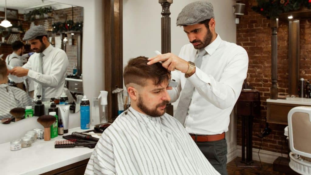 A male barber in a flat cap and tie cuts a customer's hair in a vintage-style barbershop.
