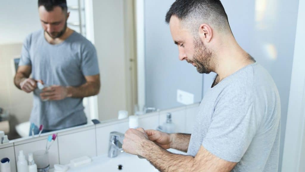 A bearded man in a gray shirt looks down, squeezing a product from a tube over a bathroom sink.