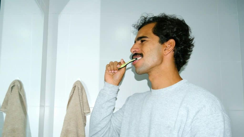 A young man with dark, curly hair and a mustache brushes his teeth in a bright bathroom.