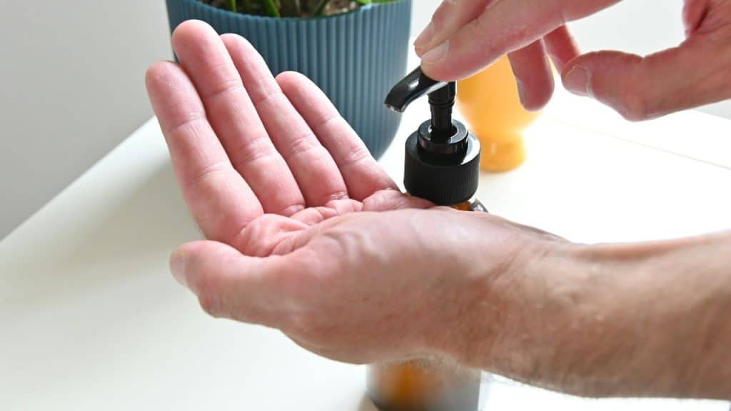 A person's hand is held under a pump bottle to receive lotion on a white counter.