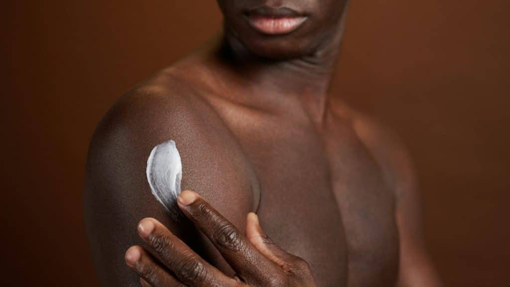 Close-up of a muscular man applying a thick streak of white moisturizer to his shoulder.