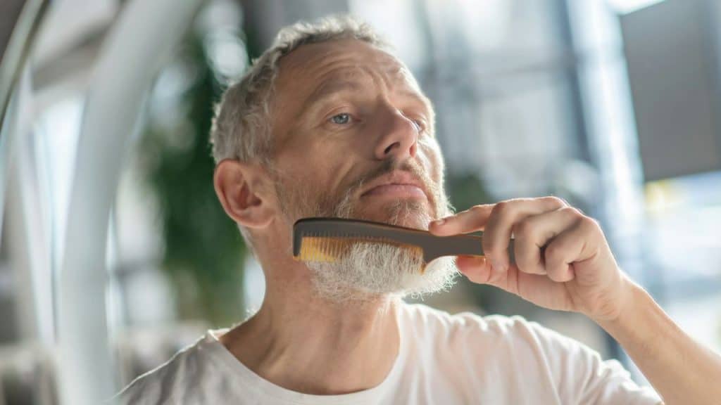 A gray-haired man in a white shirt combs his salt-and-pepper beard upward using a dark comb.