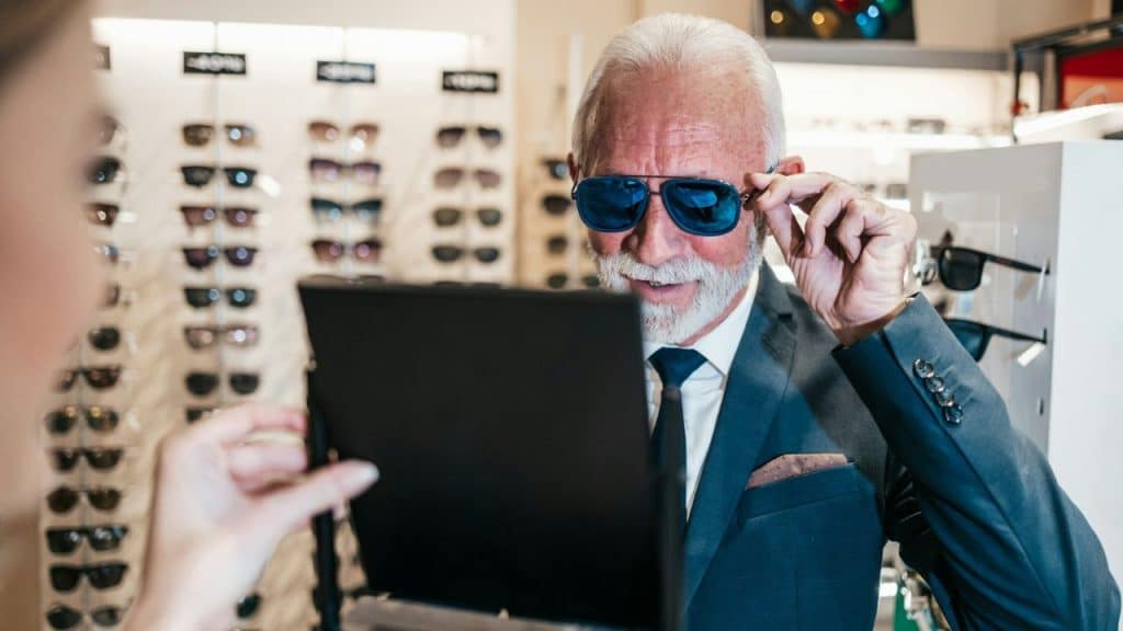 An older, well-dressed man smiles, trying on blue-mirrored sunglasses in an optical store.