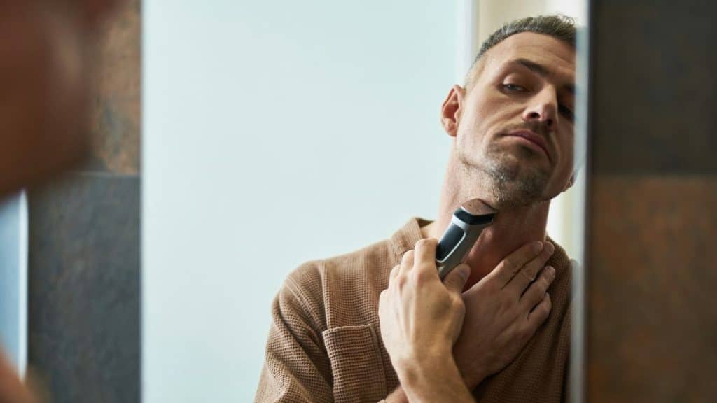 A serious man in a brown bathrobe trims the hair on his neck using an electric shaver.