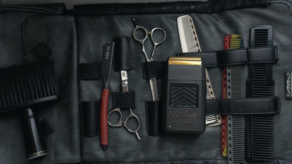 An overhead view of various barber tools, including a shaver, scissors, and combs, laid out on a black leather kit.