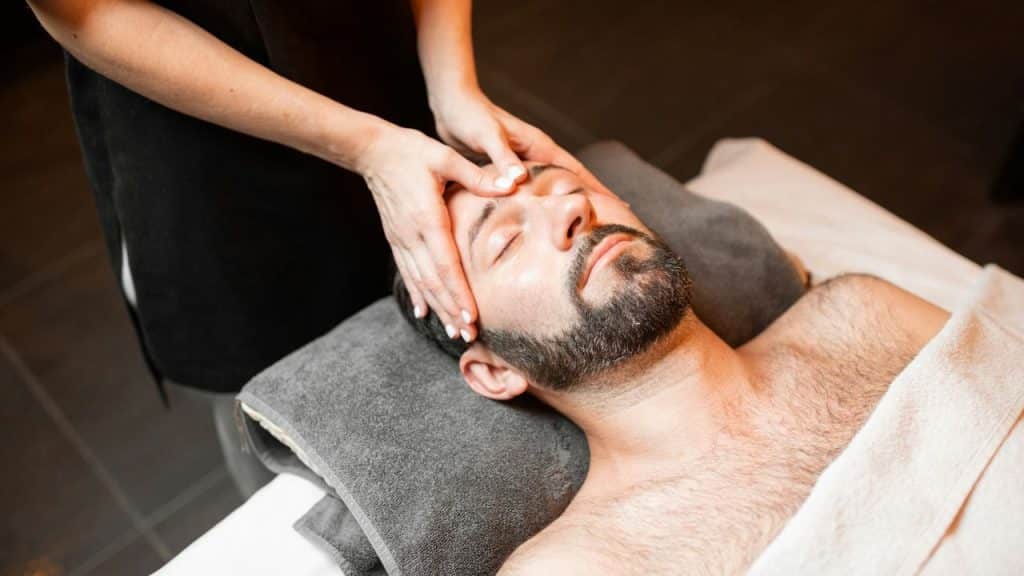 An overhead view of a bearded man with his eyes closed receiving a facial massage from a woman.