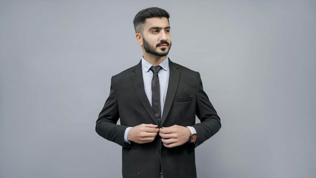 A neatly groomed, bearded man in a black suit and tie buttons his jacket