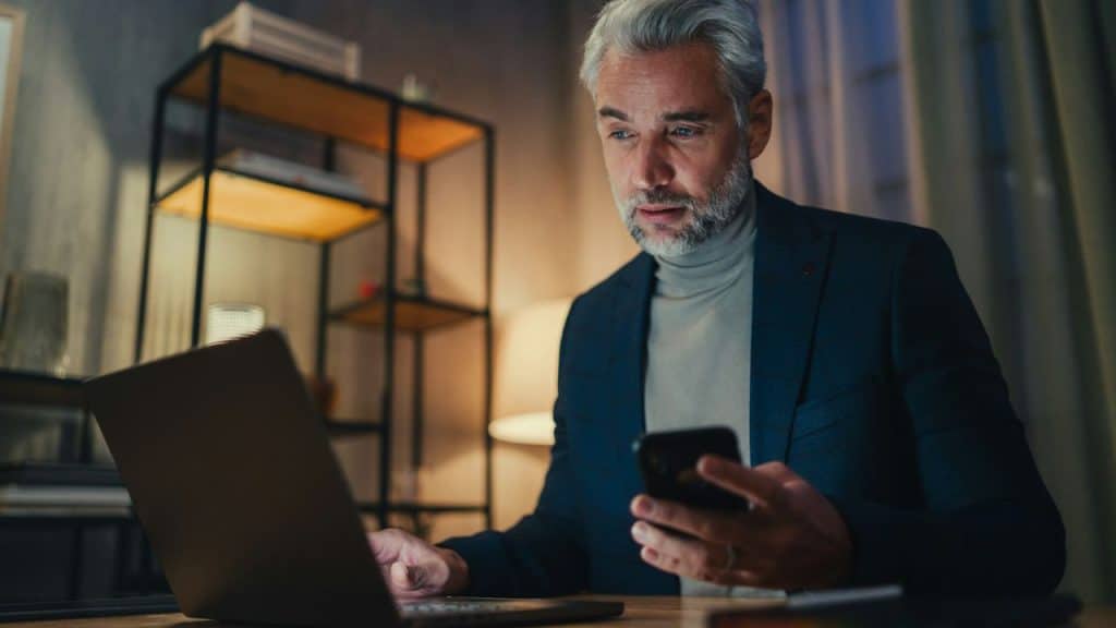 A senior man with gray hair and beard works on a laptop while holding a smartphone at night.