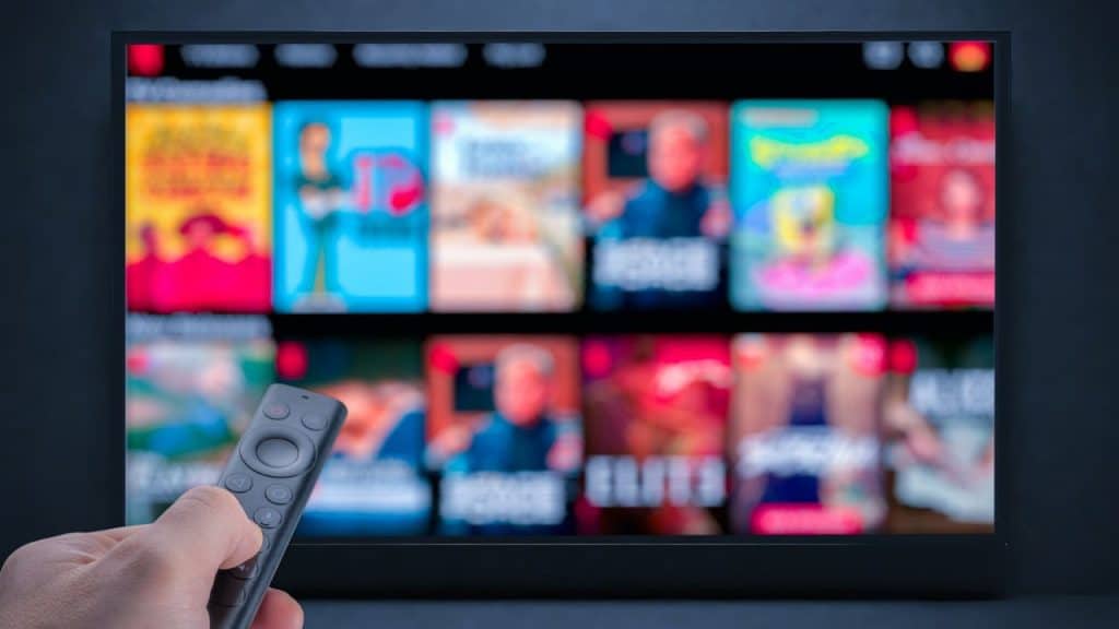 Hand holding a black remote pointing at a television screen displaying a blurry grid of colorful show thumbnails.