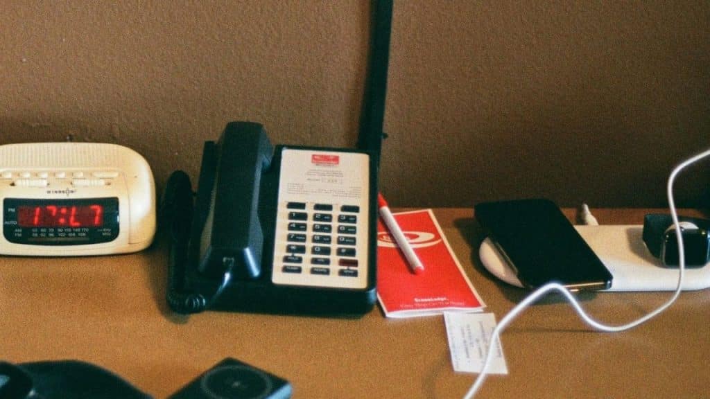 A tabletop still life showing a black landline phone, a digital clock displaying 17:47, and a smartphone on a wireless charger.
