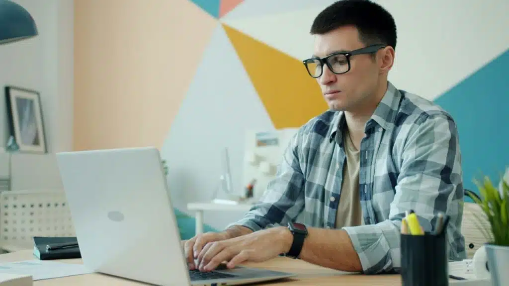 Young man wearing a plaid shirt and black-rimmed glasses intently typing on a silver laptop at a desk.