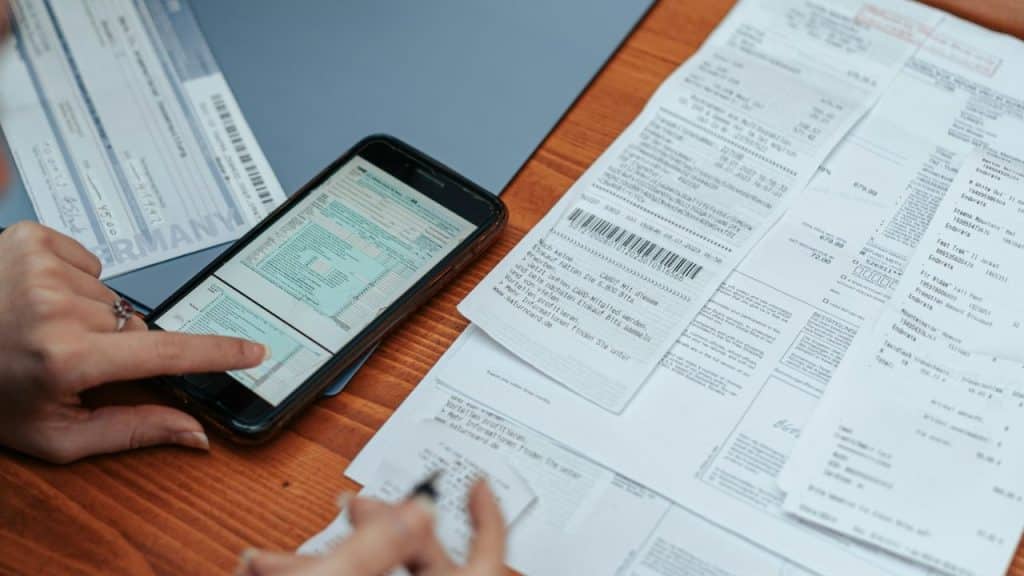 Pair of hands holding a smartphone over a messy pile of receipts and paper bills on a table.