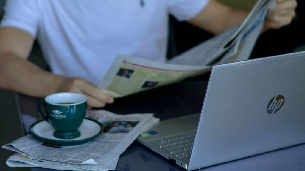 Person in a white shirt reading a newspaper next to a silver HP laptop and a green cup of coffee.