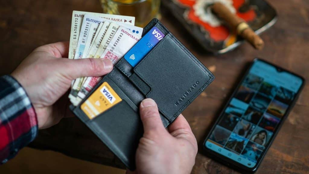 Hands holding an open black leather wallet with banknotes and VISA cards next to a smartphone showing a photo grid.