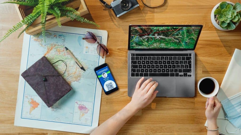 Overhead shot of a travel-themed desk setup with a world map, laptop displaying a map, a smartphone showing a COVID pass, and a cup of coffee.