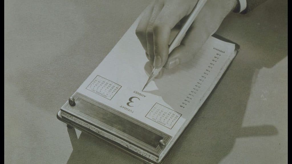 Sepia-toned, close-up shot of a hand writing on a desk calendar dated Tuesday, August 3.