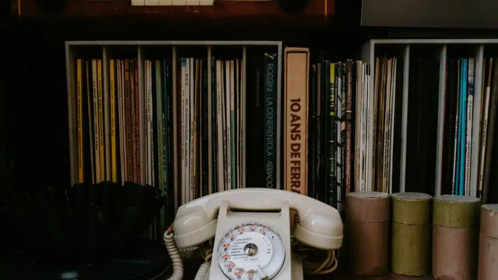 Still life of an off-white rotary telephone on a shelf surrounded by upright vinyl records and books.