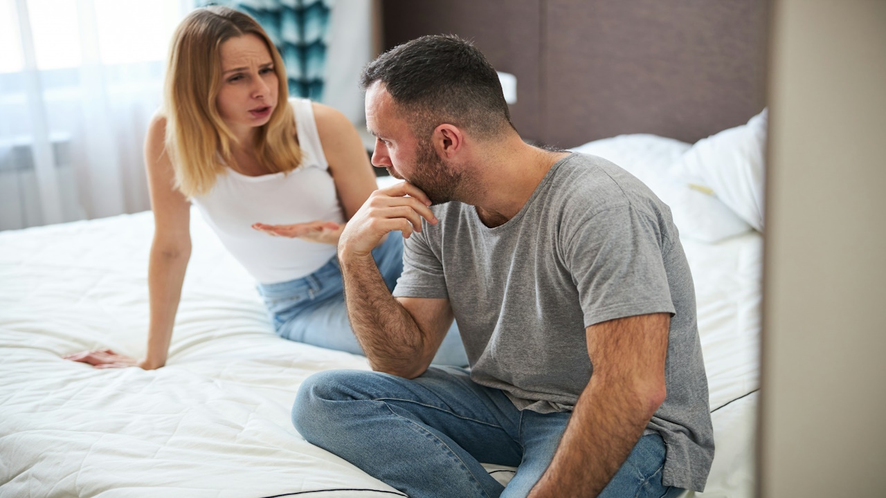 A displeased blonde lady sitting on bed and nagging her husband while the man touches his chin.