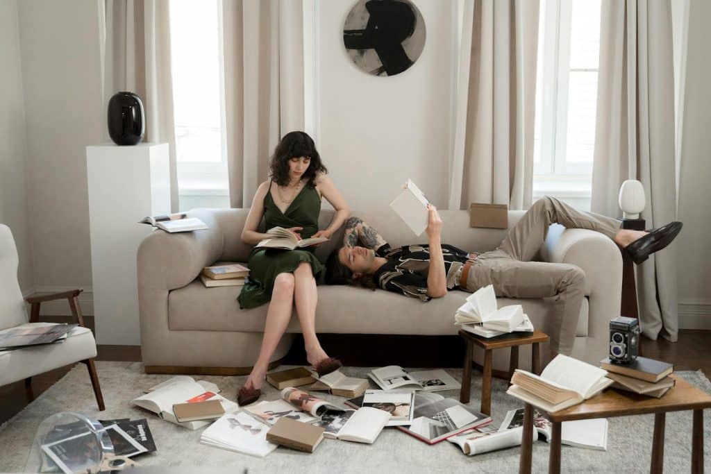 A couple sitting on their sofa reading with a lot of books scattered on the floor.