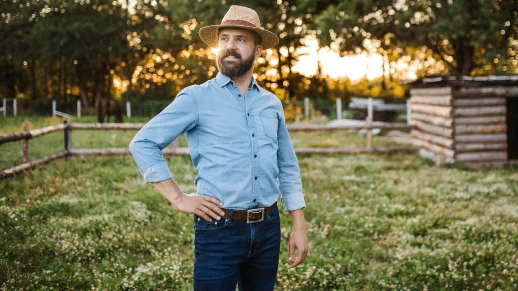 A smiling, confident man in a straw hat and denim shirt stands in a sunny field.