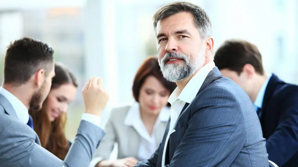 Older man with beard looks toward the camera during an office meeting.