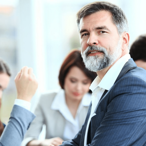 Older man with beard looks toward the camera during an office meeting.