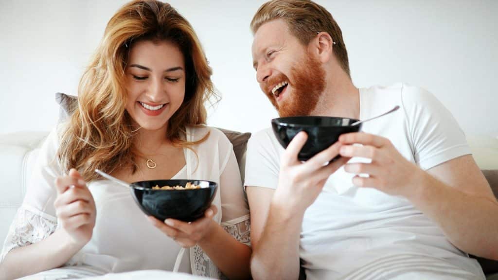 A couple having cereal in bed and laughing together.